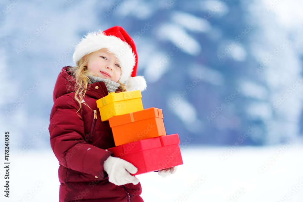 Adorable little girl wearing Santa hat holding a pile of Christmas gifts on beautiful winter day Adorable little girl wearing Santa hat holding a pile of Christmas gifts on beautiful winter day