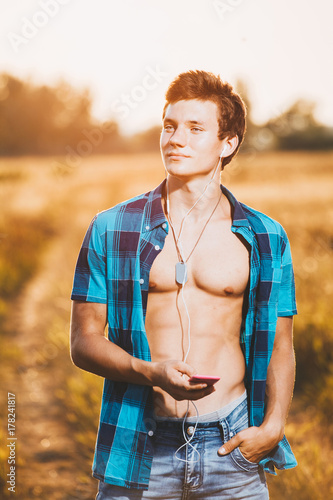 a handsome young man with muscular torso listening to music in white headphones and holding a smartphone in his hand. He is dressed in a shiny blue shirt and jeans shorts, is in a field outside city © Elizaveta