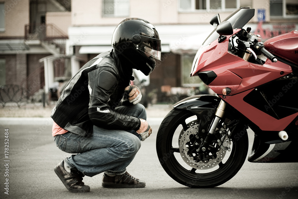 Fototapeta premium facet motocyklista w kasku i skórzanej kurtce i dżinsach siedzi naprzeciw motocykla sportowego czerwonego koloru i patrzy na niego twarzą w twarz