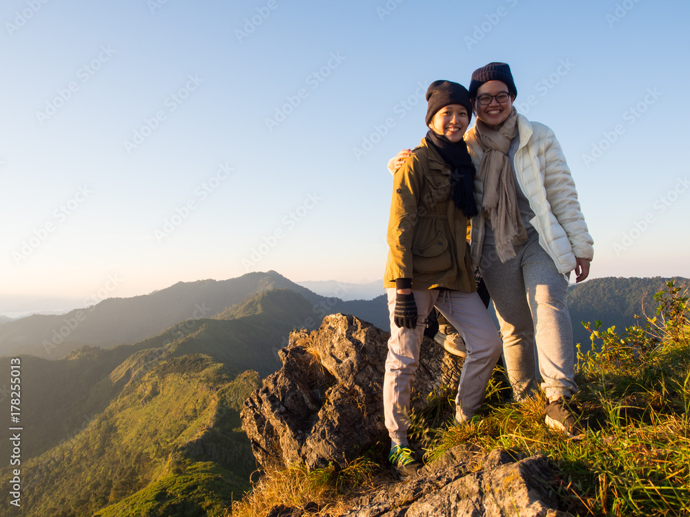 Two best friends smile happily on the top among the range of mountain ridge with yellow sunlight of morning sunrise. The seek of true friendship can be found from trekking at Doi Phu Wae, Nan Thailand