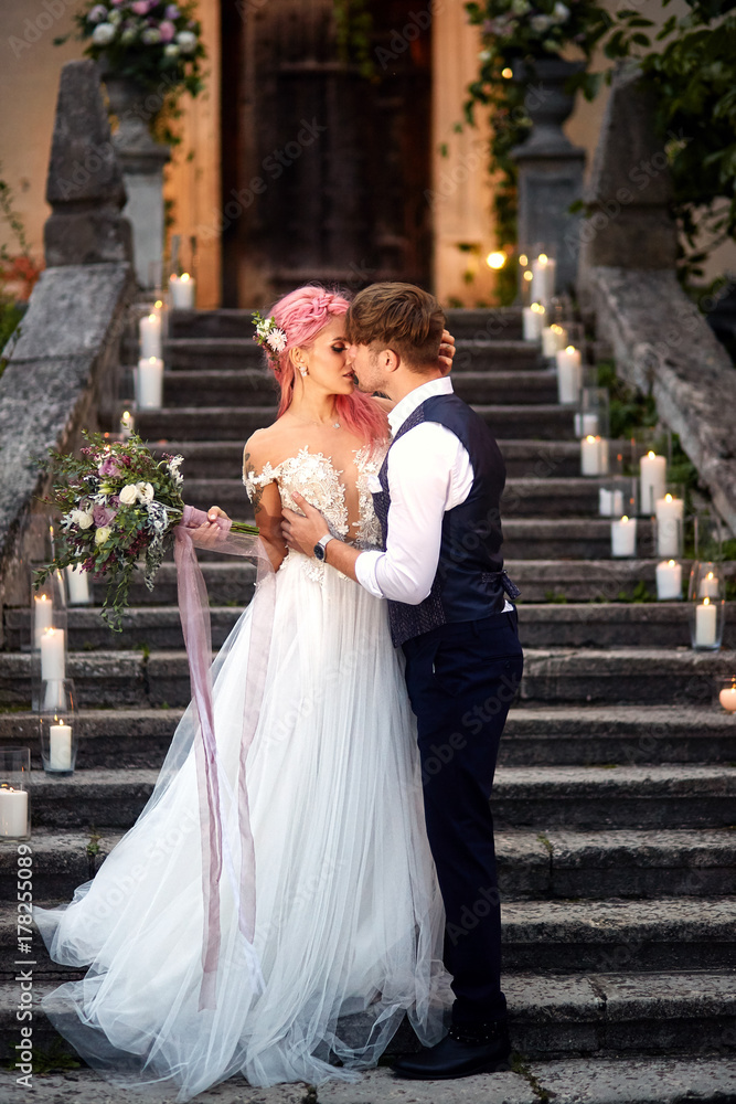 Bride with pink hair and stylish groom stand on footsteps with shiny candles