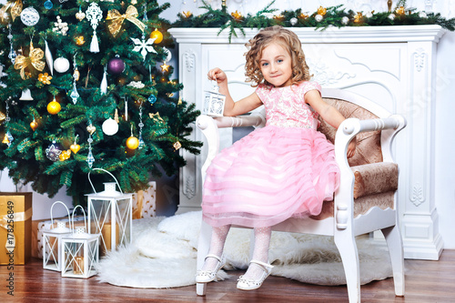 little cute girl in a pink dress is sitting in a room by the fireplace and Christmas tree