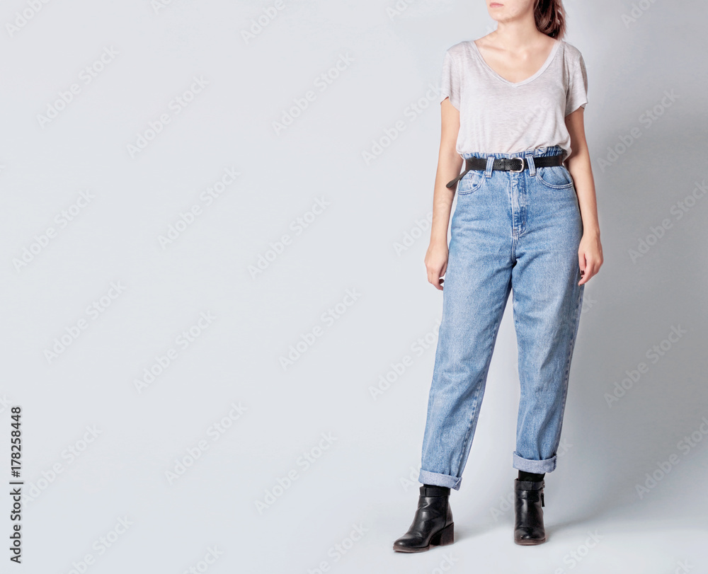 Woman wearing casual outfit with white t-shirt, blue vintage high-waisted  mom jeans with black faux leather ankle boots and black belt isolated on  grey background. Copy space Stock Photo