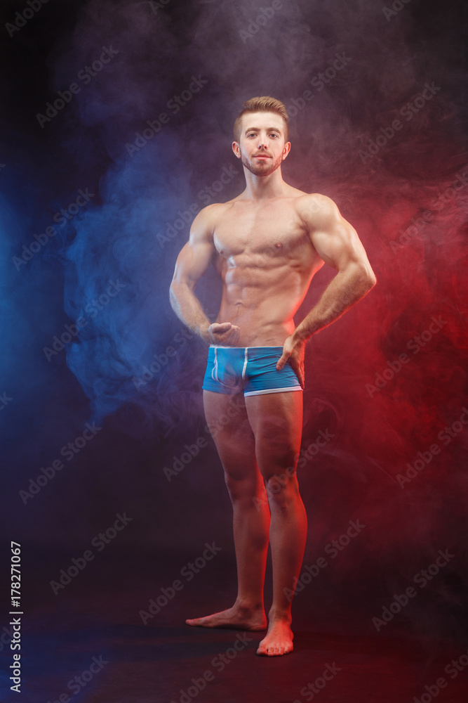 Bodybuilding, sports, weightlifting, fitness concept. Young muscular fit man with perfect body posing at dramatic smoky background, vertical image