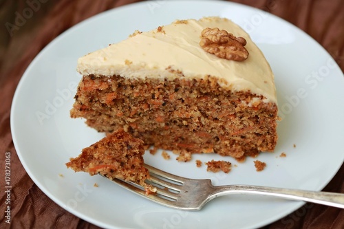 Angle view of a piece of homemade carrot cake with cream cheese topping decorated with a piece of walnut placed on white plate with silver fork put on brown tablecloth