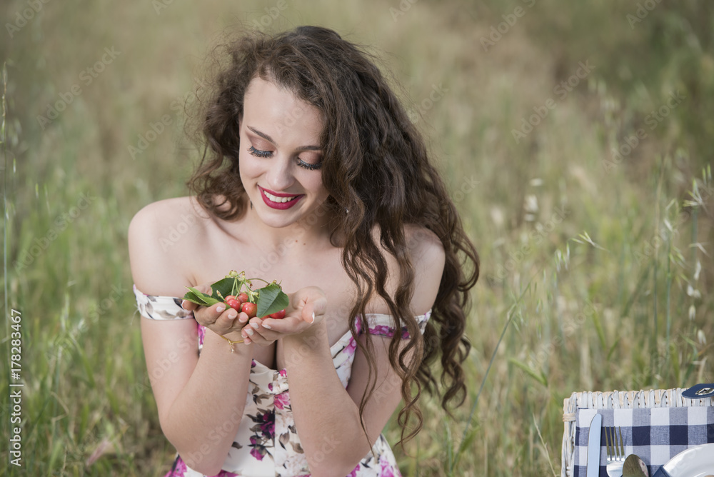 Sweat young woman sitting in dry field wearing a floral short dress holding some cherries in her hands, open rerto picnic basket at her side Sweat young woman sitting in dry field wearing a floral short dress holding some cherries in her hands, open rerto picnic basket at her side