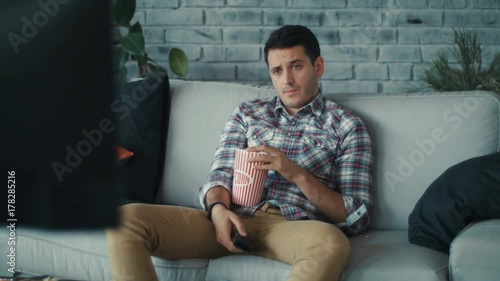 Close up young man watching the TV siting on the sofa with remote in his hands use phone in home handsome laziness looking television men shot on RED EPIC camera. Slow Motion Shot.