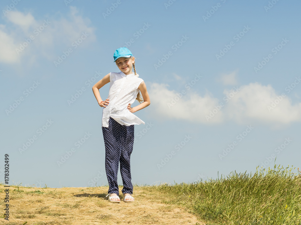 Beautiful happy girl in a blue baseball cap on top of a hill on a background of blue sky with clouds.