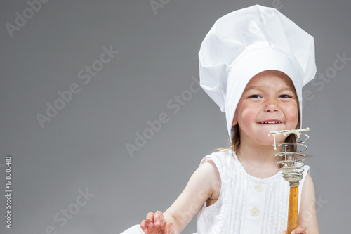 Funny Cooking Concepts. Portrait of Smiling Little Caucasian Girl in Cook Uniform Posing with Whisk Covered with Flour. Against Gray. photo