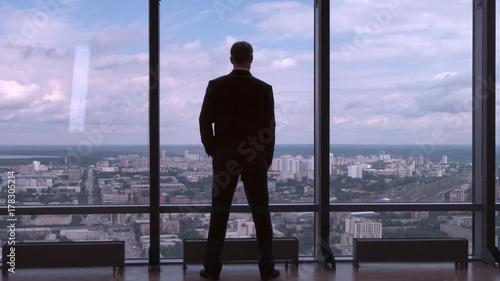 Full length back view of successful businessman in suit standing in office with hands on waist, CEO. Businessman from the back in front of a city view on the window