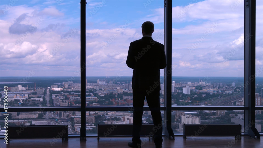 Businessman in suit standing next to a big window from an office building and checking his watch back view. Businessman standing at window looking on his wrist watch