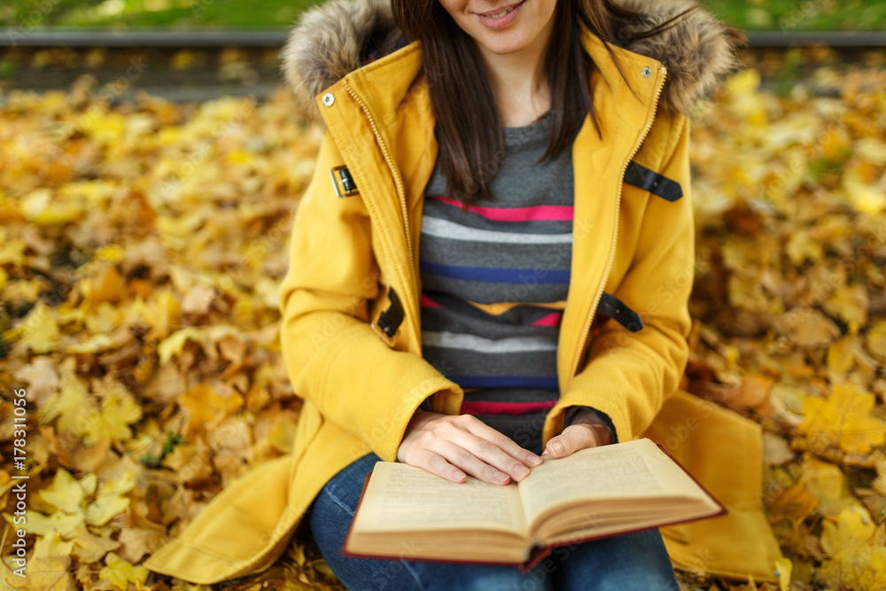 Woman in yellow coat and jeans sitting under the maple tree with a red book in fall city park on a warm day. Autumn golden leaves. Reading concept. Close up. Woman in yellow coat and jeans sitting under the maple tree with a red book in fall city park on a warm day. Autumn golden leaves. Reading concept. Close up.