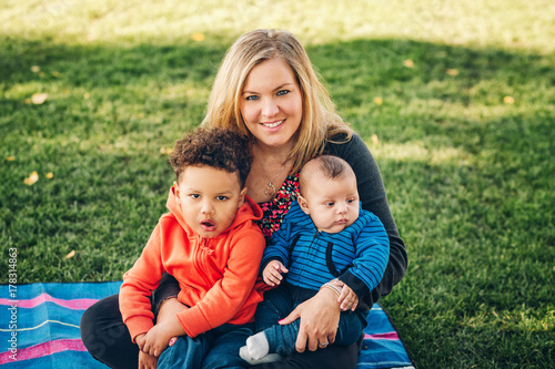 Happy family resting in the park on a nice sunny day, young mother playing with two little sons outside. Baby and toddler boys holding by loving parent