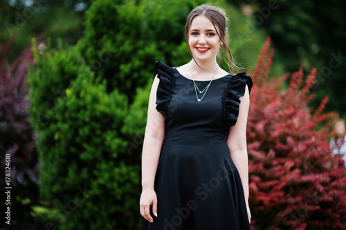 Portrait of a gorgeous young girl in black dress walking on the pavement in the park on a prom day.