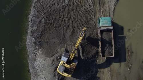 Yellow earth mover collecting sand with hydraulic shovel and loading tipper by the river on sandbank, aerial view, extreme high angle, top shot,  drone flying up from digger and dump truck, sunny day