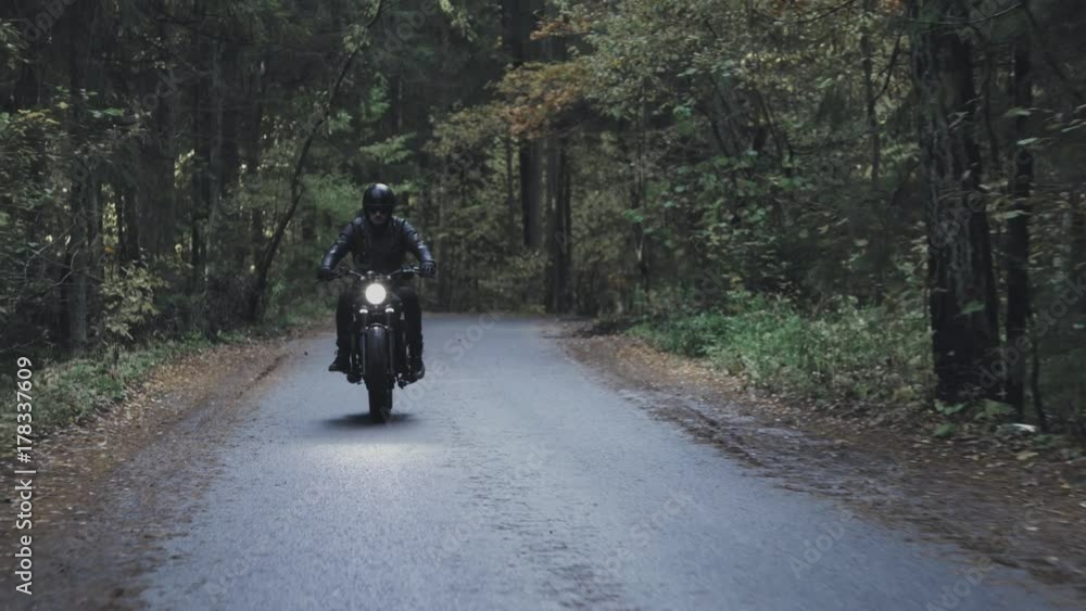 A guy in a black leather jacket and helmet riding a classic motorcycle on a forest road. A guy in a black leather jacket and helmet riding a classic motorcycle on a forest road.