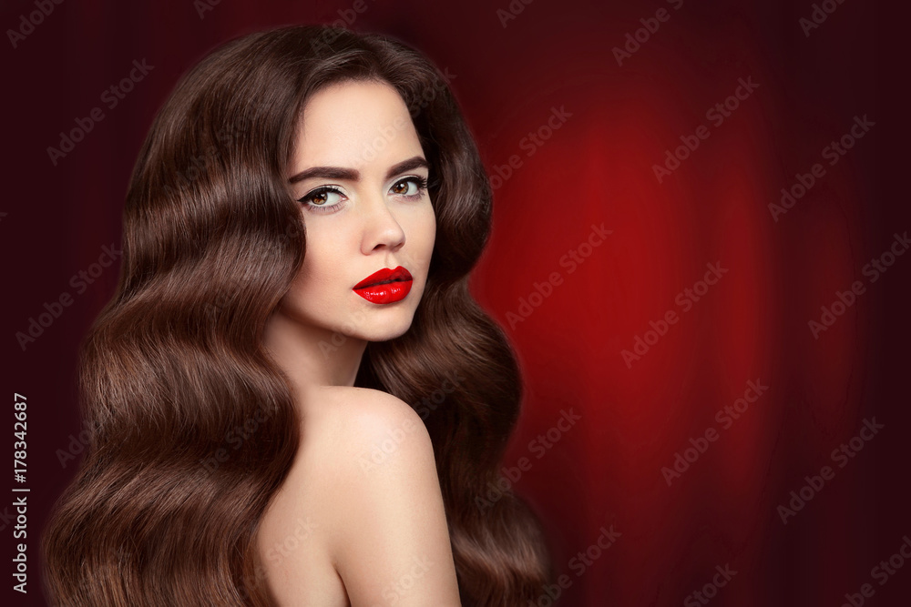 Fototapeta premium Beauty hair. Brunette girl portrait with red lips makeup and long shiny wavy hair. Beautiful model with healthy hairstyle isolated on drapery curtains dark background. Gorgeous Glamour Lady.