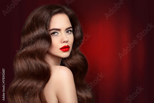 Beauty hair. Brunette girl portrait with red lips makeup and long shiny wavy hair. Beautiful model with healthy hairstyle isolated on drapery curtains dark background.  Gorgeous Glamour Lady.