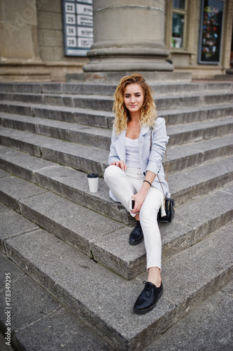 Stylish curly blonde model girl wear on white with cup of coffee posing on stairs outdoor.