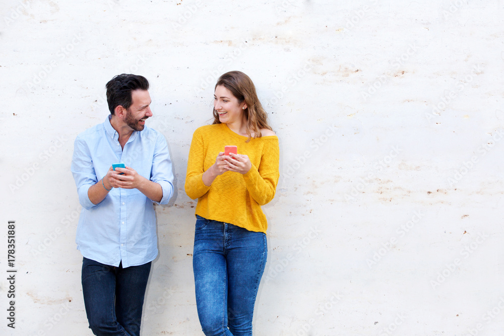 Laughing couple standing by white wall holding mobile phones