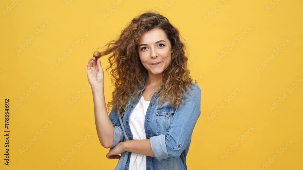 Smiling mystery curly woman in denim shirt playing with her hair and looking at the camera over yellow background Smiling mystery curly woman in denim shirt playing with her hair and looking at the camera over yellow background