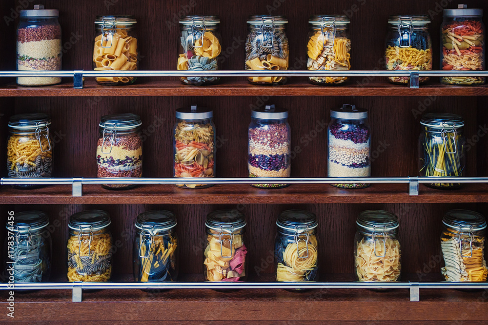 On the wooden shelves are glass jars with various varicoloured types of pasta, spaghetti, beans, cereals for their storage and decorating the kitchen