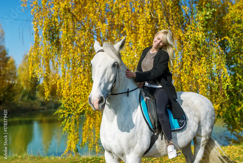Young woman riding a horse on a sunny autumn day against the background of the blue lake