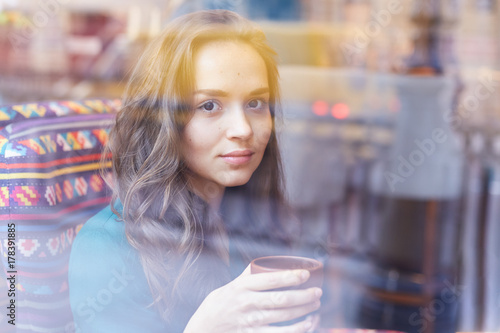 Attractive stunning young lady sitting alone at cafe table, captured through the window. Beautiful Caucasian woman relaxing on couch at modern restaurant, drinking tea, looking at camera with smile.