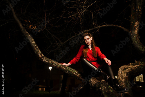 Black girl in red shirt and bright make-up on night street sitting on scary tree outdoor. Halloween theme.