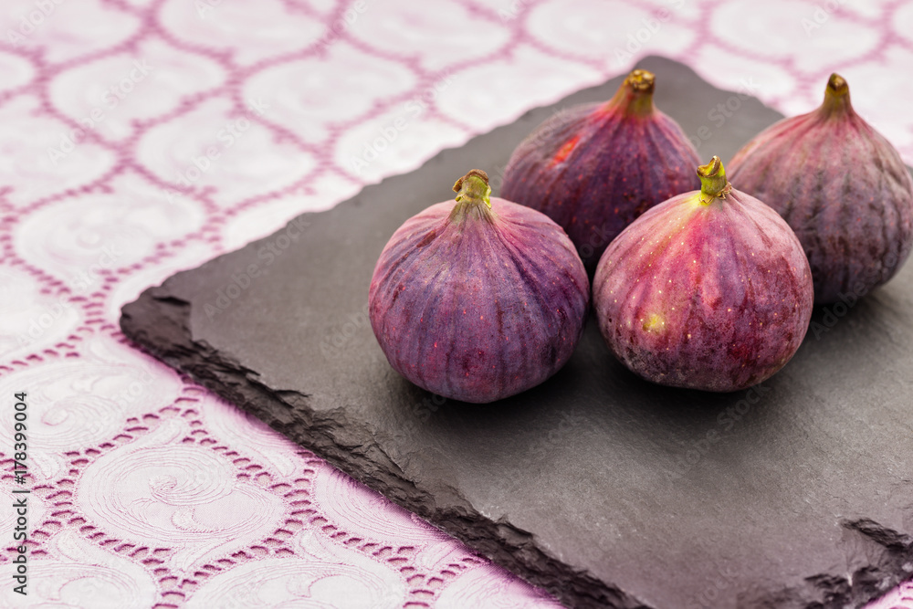 Fototapeta premium Four fresh purple Figы on black slate plate on purple lace tablecloth 