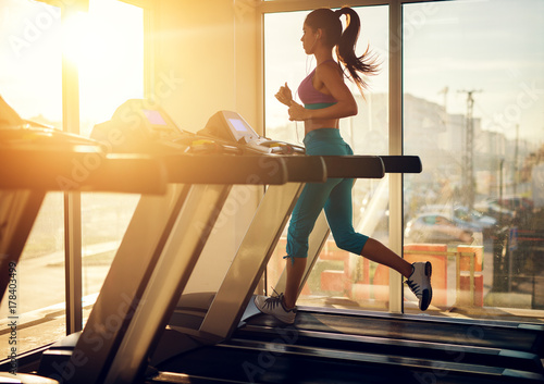 Canvas Print Young healthy athletic woman running on a treadmill near the sunny window in the gym and listening music