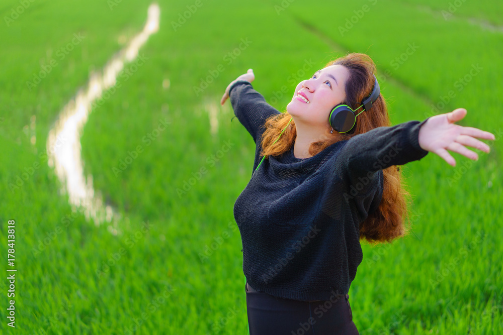 Happy Asian girl raising arms is listening to music with headphone. She is in paddy field of Thailand.