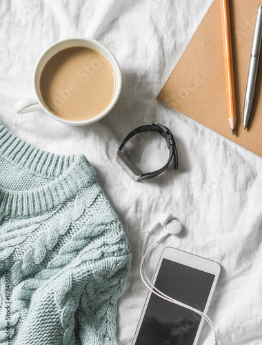 Blue knitted sweater, coffee with milk, notebook, headphones, smart phone on bed, top view. Women clothing. Flat lay