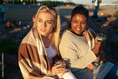 Intercultural girls with drinks admiring sunset on the beach on summer evening