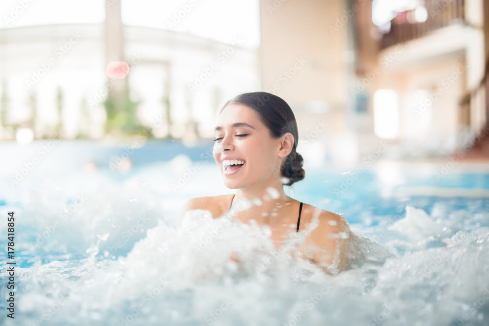 Excited female laughing while splashing warm water during spa procedure in whirlpool - obrazy, fototapety, plakaty 