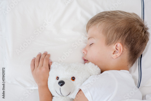 Little boy sleeping in bed with toy bear. Top view