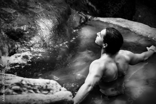  Нandsome guy with long hair and piercings on waterfalls in a rain forest against a background of clear blue and green water. Tarzan concept. Black and white photo.