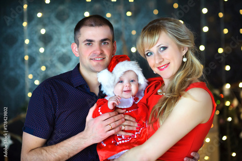 Obraz na płótnie happy family with a baby in a Santa Claus costume hugs against a bright festive lights. w salonie