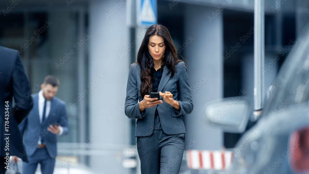 Obraz premium Business Woman in the Tailored Suit Walking on the Busy Big City Street in the Business District, Checks Her Smartphone. Confident Woman on Her Way to do Big Business.