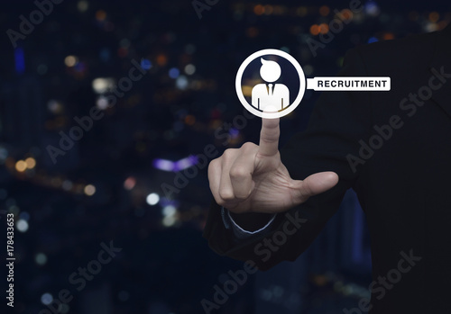 Hand pressing businessman with magnifying glass icon over blur colorful night light city tower, Business recruitment concept