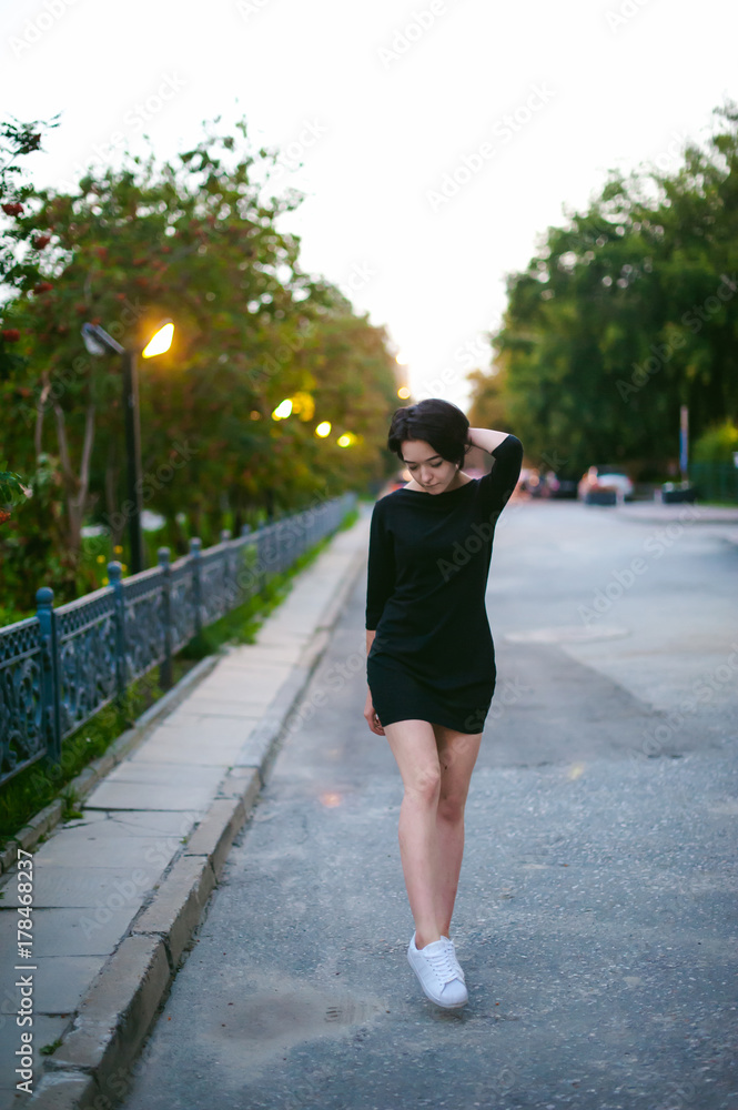 young woman with beautiful feet dressed in white sneakers and black dress, against a backdrop of urban landscapes. Street fashion. dark style