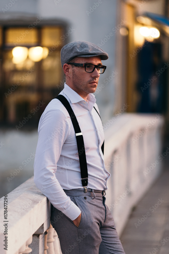 Fashionable retro dressed man with cap, suspenders and eyeglasses standing on city street and looking at side.