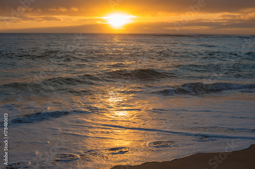 Shorebreak foam, ocean water lit with sunset light, warm clouds on background