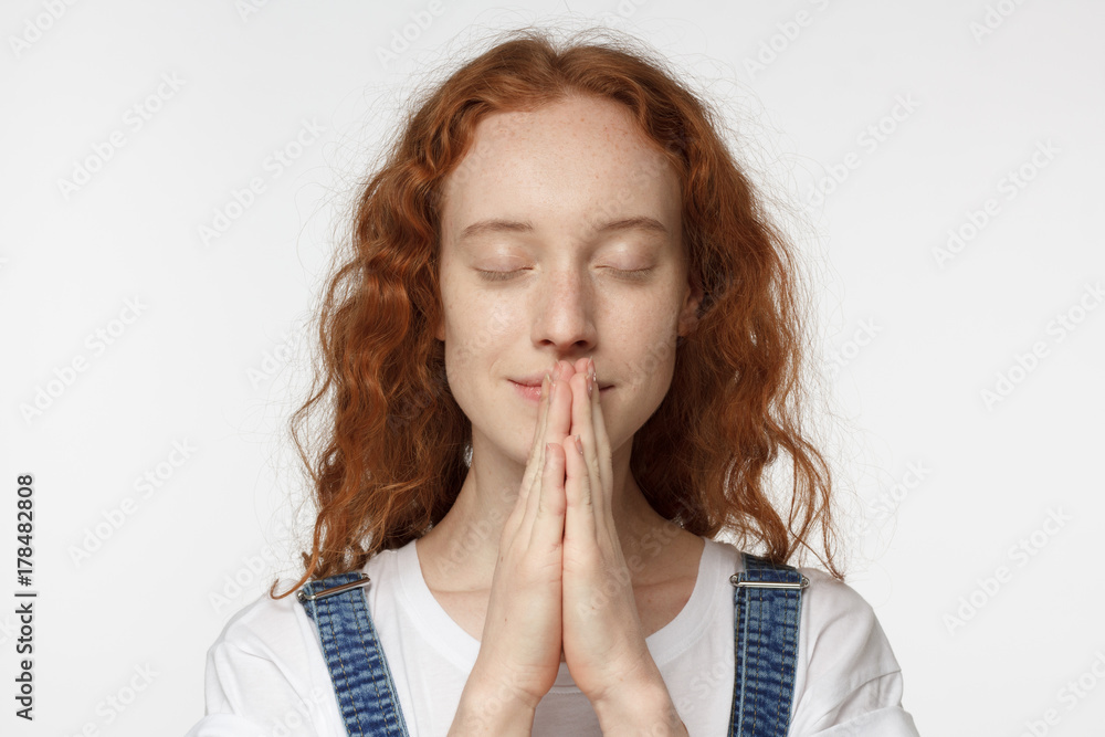 Closeup of curly redhead girl isolated on gray background putting hands together as if she is praying with closed eyes, smiling Closeup of curly redhead girl isolated on gray background putting hands together as if she is praying with closed eyes, smiling