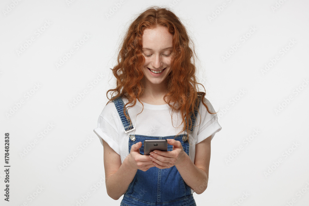 Indoor shot of good-looking young redhead female isolated on gray background looking at smartphone, smiling openly while holding smartphone in both hands Indoor shot of good-looking young redhead female isolated on gray background looking at smartphone, smiling openly while holding smartphone in both hands