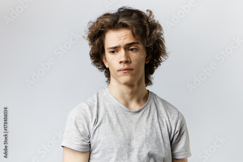 Worried young male with curly hair pondering at blank grey wall. Frustrated 20 year old guy having thoughtful pensive expression on his smooth face, worrying about his future life and career