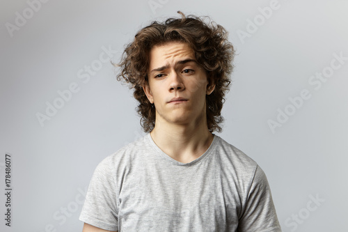 Isolated portrait of puzzled young male with curly hair frowning eyebrows and biting lips, worrying about bad grades or problems at university, having deep in thoughts pensive facial expression