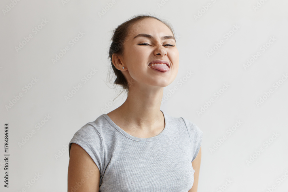 Naughty girl misbehaving, sticking out her tongue at camera as a sign of disobedience, protest and disrespect. Picture of Caucassian young woman grimacing and having fun at blank studio wall Naughty girl misbehaving, sticking out her tongue at camera as a sign of disobedience, protest and disrespect. Picture of Caucassian young woman grimacing and having fun at blank studio wall