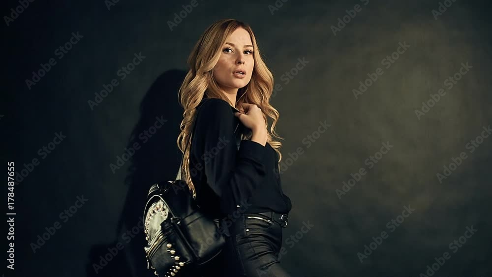 Sensual portrait of beautiful blonde woman with curly gold hair in dark clothes and leather bag posing on black background in studio Sensual portrait of beautiful blonde woman with curly gold hair in dark clothes and leather bag posing on black background in studio