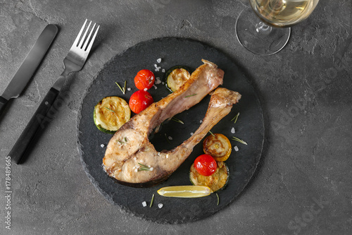Slate plate with fried fish steak and vegetables on grey background